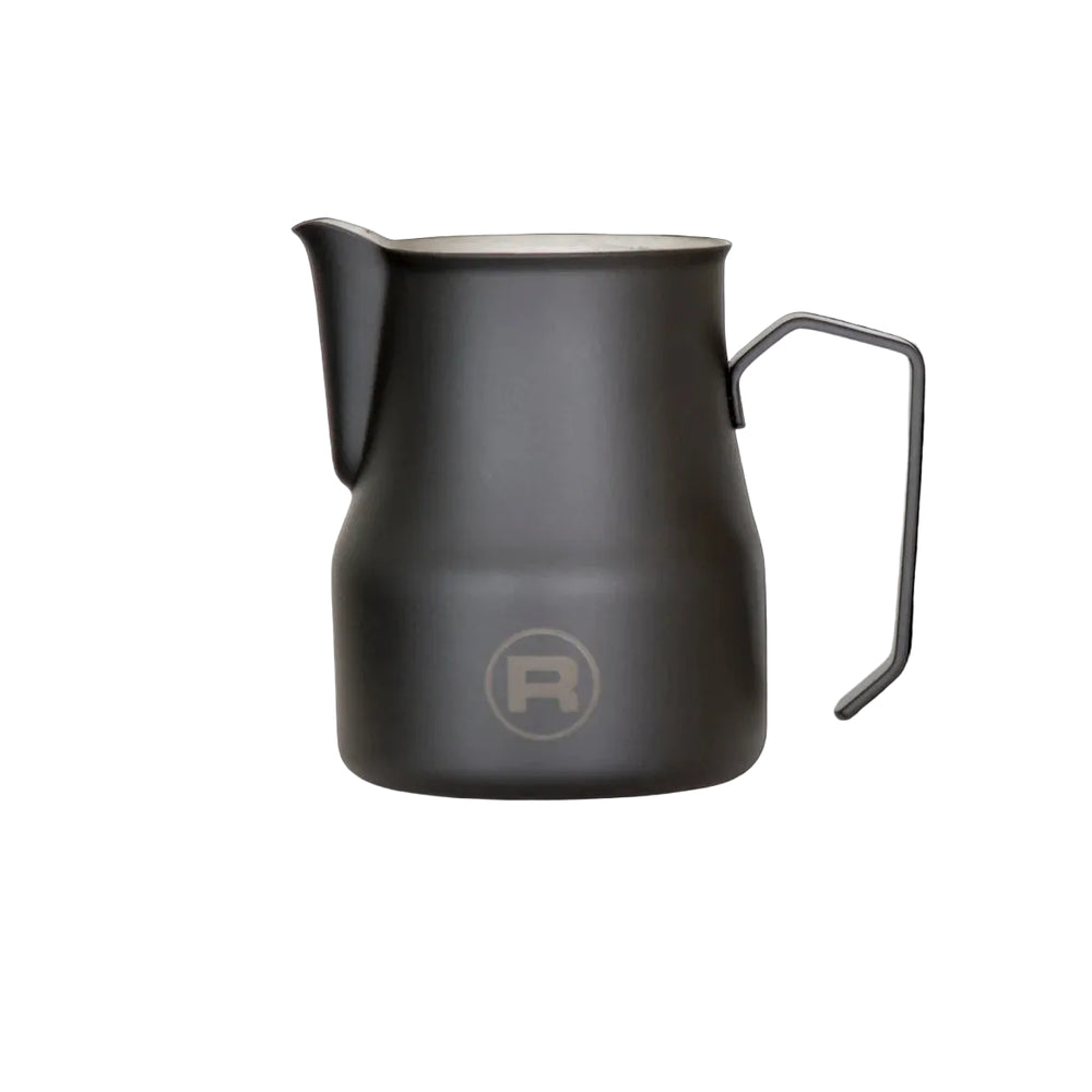A matte black metal milk pitcher with a handle and a frosted glass spout, featuring a circular 'R' logo on the front, set against a solid black background.